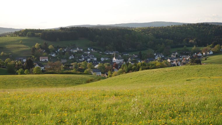 Eine malerische Aussicht auf ein Dorf in einer grünen Landschaft. Sanfte Hügel und blühende Wiesen umgeben die kleine Ortschaft.