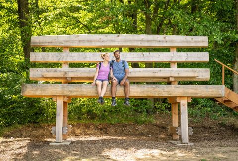 Ein großes Holzgestell im Freien, auf dem ein Paar sitzt. Es ist von grünen Bäumen umgeben und strahlt eine entspannte Atmosphäre aus.