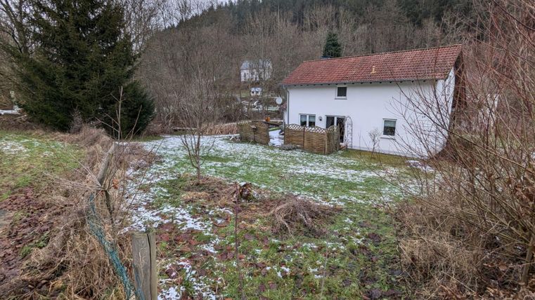 A simple house in a rural area, surrounded by trees and a winter landscape. The ground is partly covered with snow.