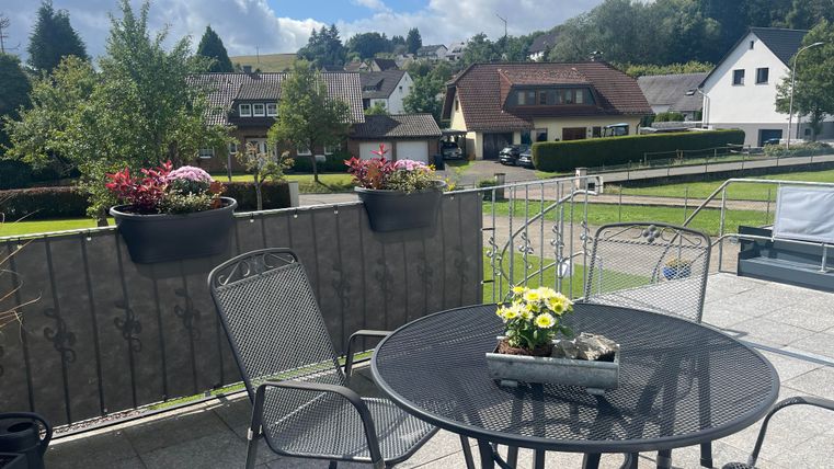 Eine einladende Terrasse mit einem runden Tisch und Stühlen. Im Hintergrund sind grüne Bäume und einige Häuser zu sehen.