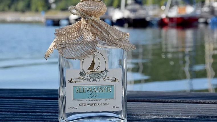 Eine Flasche Seewasser-Gin steht auf einem Holzsteg am Wasser. Im Hintergrund sind Boote und eine grüne Landschaft zu sehen.