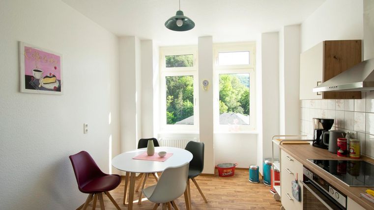 Eine helle Küche mit einem runden Tisch und bunten Stühlen. Große Fenster lassen viel Tageslicht herein und bieten einen schönen Ausblick auf die Natur.