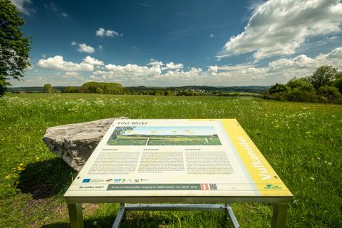 Im Vordergrund steht mitten auf einer blühenden Wiese ,eine Informationstafel zum Eifelblick Steinbüschel.Links liegt ein großer Stein.Weit im  Hintergrund sieht man den Ort Schüller auf einer Anhöhe liegen. Es ist ein sonniger Tag mit blauem Himmel und einigen Wolken. Die Fernsicht ist so gut, dass man iin der Ferne sogar den Aremberg erkennen kann.