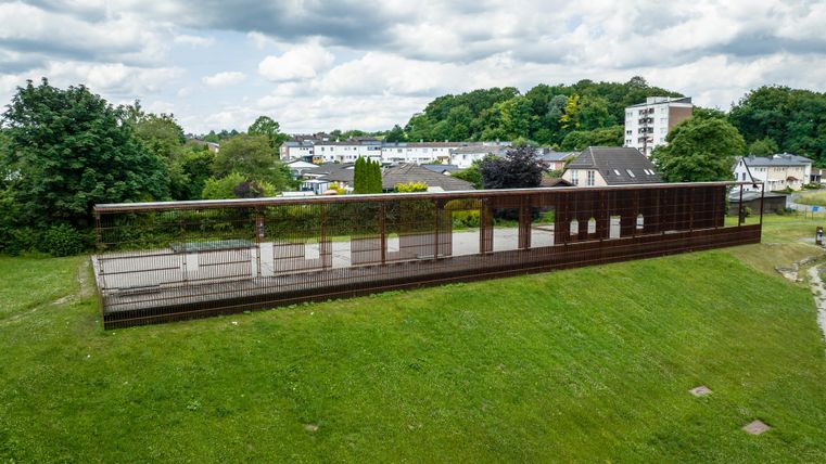 A modern observation platform made of metal stands on a green hill. In the background, trees and buildings are visible under a cloudy sky.