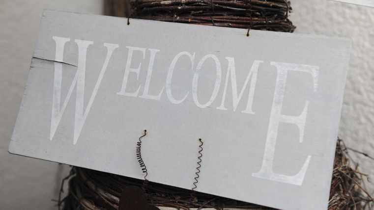 Ein Willkommensschild aus Holz in neutralen Farben. Es hängt an einer dekorativen Struktur und vermittelt eine freundliche Atmosphäre.