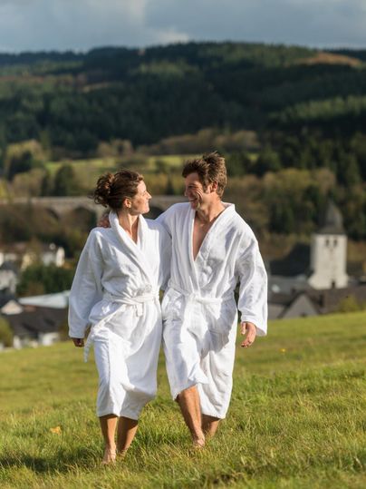 Ein Paar in weißen Bademänteln spaziert entspannt über eine Wiese. Im Hintergrund sind sanfte Hügel und ein kleines Dorf zu sehen.