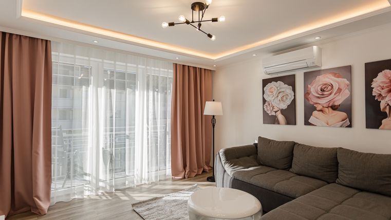 Ein modernes Wohnzimmer mit einem grauen Sofa und einem runden Tisch. An der Wand hängen Bilder von Rosen, und große Fenster lassen viel Licht herein.