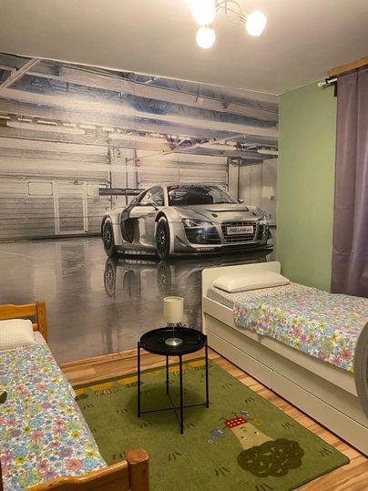 Ein modernes Schlafzimmer mit zwei Betten und einem Teppich. An der Wand ist ein großes Bild eines silbernen Sportwagens in einer Garage zu sehen.