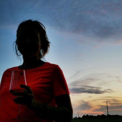 Een persoon houdt een glas in de hand, gesilhouetteerd tegen een kleurrijke avondlucht. De kleuren van de lucht variëren van blauw tot oranje.