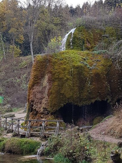 Een kleine waterval stroomt over mosbedekte rotsen. Op de voorgrond is een rustige rivier en een houten brug te zien.