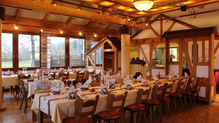 Ein geschmackvoll eingerichtetes Restaurant mit einer langen Tafel und warmem Holzdekor. Große Fenster sorgen für viel Tageslicht und eine einladende Atmosphäre.