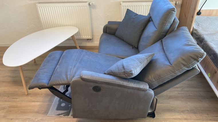 Ein bequemer, grauer Sessel steht neben einem kleinen weißen Tisch in einem hellen Raum. Der Sessel ist teilweise zurückgelehnt und hat ein Kissen.