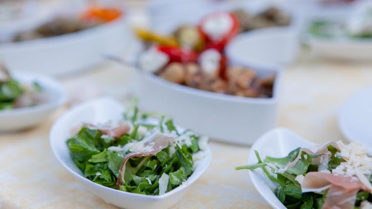 Eine Auswahl an Salaten in weißen Schalen. Im Vordergrund ist ein Rucola-Salat mit Schinken und Käse zu sehen.