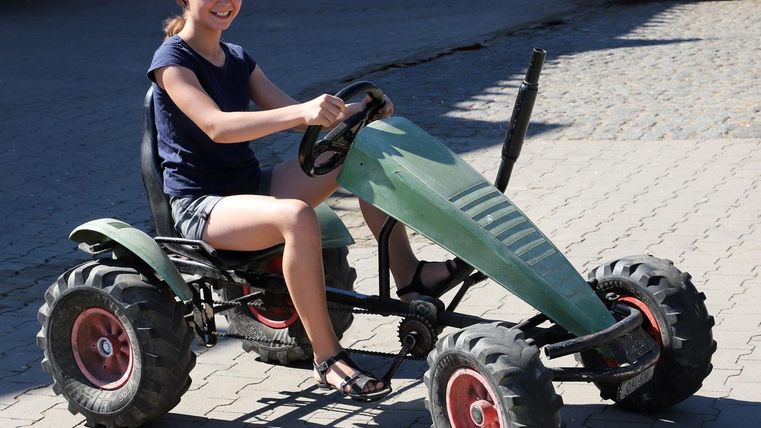 A girl is sitting on a green pedal go-kart. It is sunny and the surroundings are friendly.