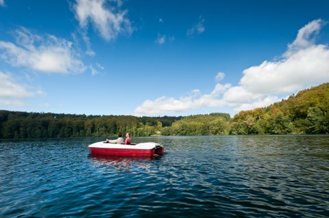 Eine Person entspannt auf einem roten Boot auf einem ruhigen See. Im Hintergrund sind Bäume und ein blauer Himmel mit einigen Wolken zu sehen.