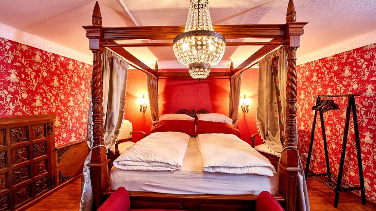 An elegant bedroom with a four-poster bed and a large ceiling light. The walls are decorated with striking red floral wallpaper.