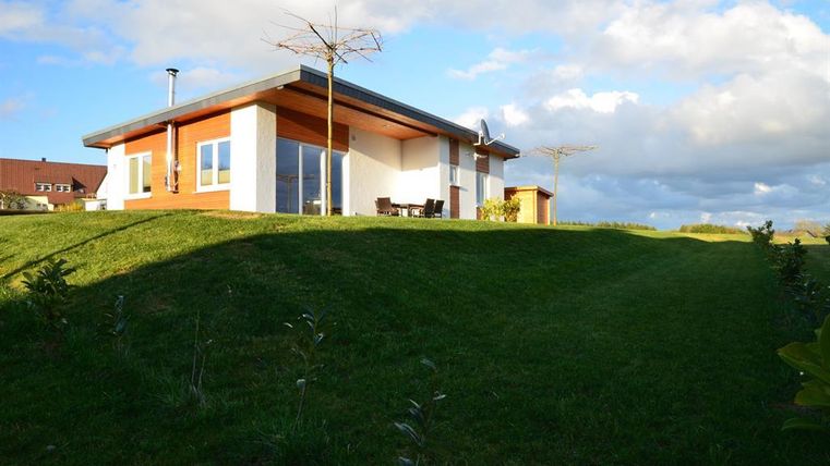 A modern house on a gentle elevation with green grass. The sky is partially cloudy and some furniture is visible on the terrace.
