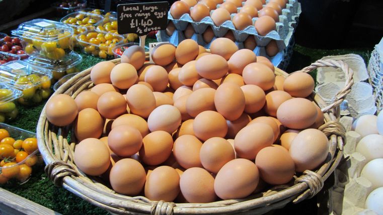 Een mand vol verse eieren ligt op een markt. Op de achtergrond zijn kleurrijke groenten en fruitsoorten te zien.