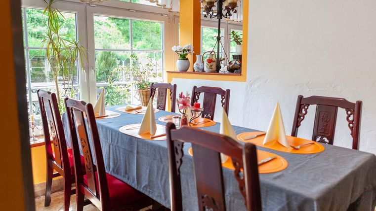 Ein stilvoll gedeckter Tisch in einem Restaurant mit Holzstühlen, grauer Tischdecke und orangefarbenen Platzsets. Pflanzen und Dekoration im Hintergrund.