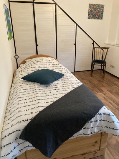 Een eenvoudig slaapkamer met een bed, een deken en een kussen. Op de achtergrond staat een stoel en een lichte trap.