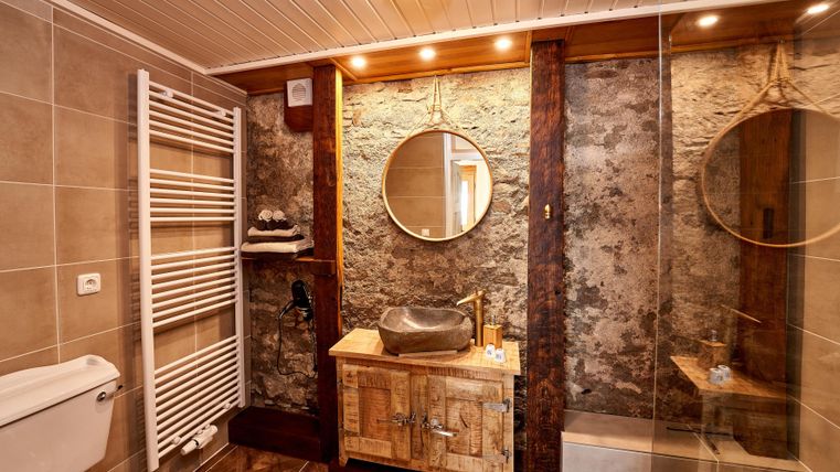 A modern bathroom with wood and stone elements. It is well-lit and features a round mirror as well as a stylish sink unit.