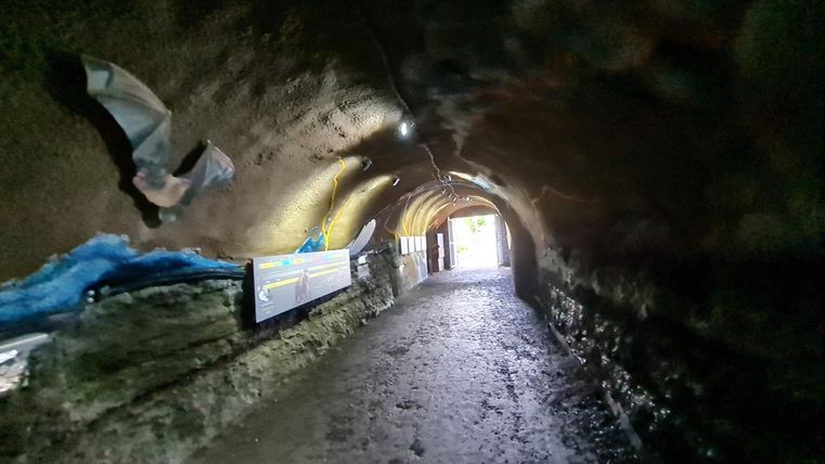 Een lange, gebogen tunnel met vochtige wanden en een heldere uitgang aan het einde. Aan de zijkanten zijn enkele borden bevestigd.