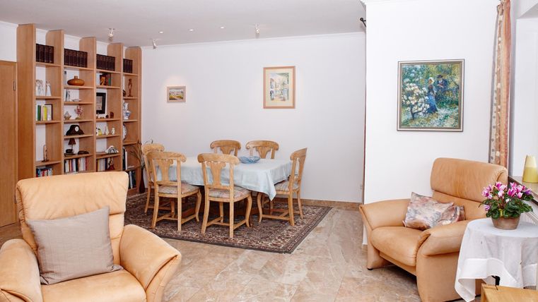 Ein helles Wohnzimmer mit gemütlichen Sessel und einem Esstisch. Im Hintergrund sind Bücherregale und Wandbilder zu sehen.