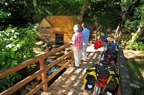 Two people are standing on a wooden bridge, looking at a small stream. Next to them are bicycles with luggage.