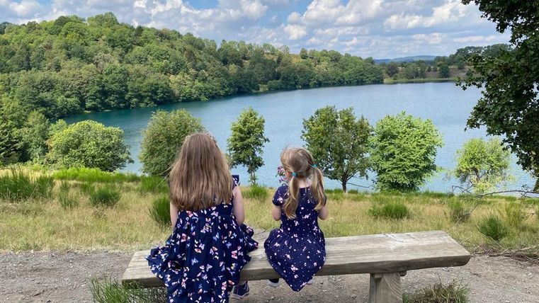 Zwei Mädchen sitzen auf einer Bank am Ufer eines Sees. Im Hintergrund sind grüne Bäume und ein blauer Himmel zu sehen.
