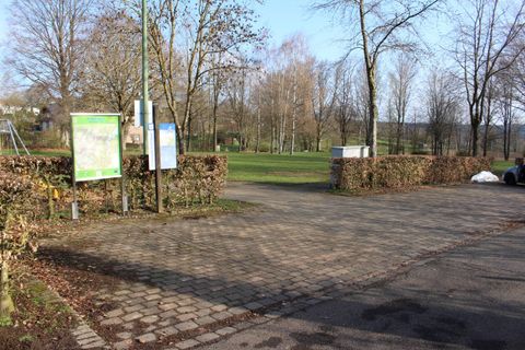 Een parkeerplaats die aan een park grenst, met banken en borden. Op de achtergrond zijn bomen en een heldere lucht te zien.