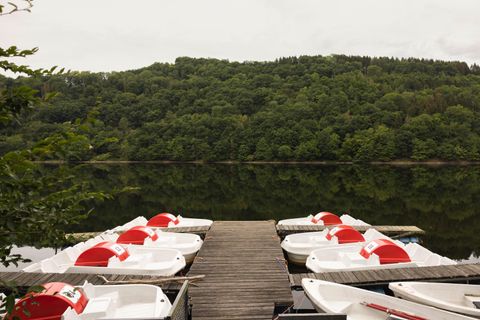Ein ruhiger See mit einem Steg, der mit Tretbooten ausgestattet ist. Im Hintergrund sind grüne Hügel und eine bewölkte Himmelsstimmung zu sehen.