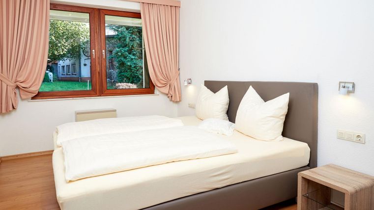 Een gezellige slaapkamer met een tweepersoonsbed en grote, witte kussens. Vanuit het raam zie je een groene tuin.