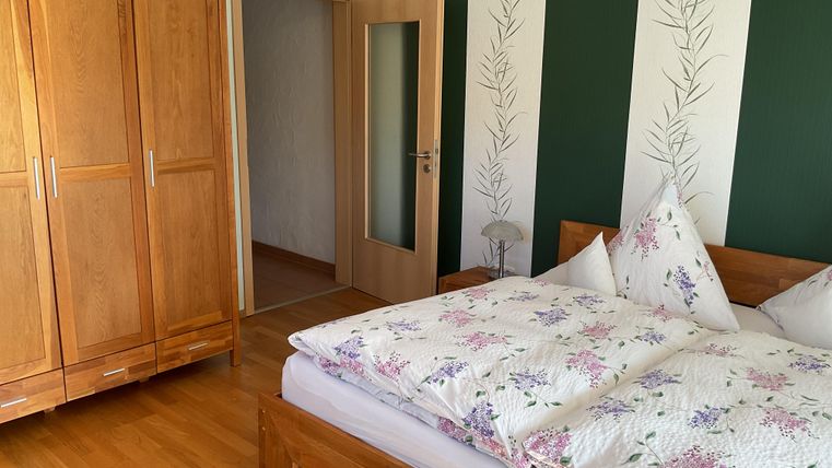 A cozy bedroom with a wooden wardrobe and a double bed. The walls are painted green and decorated with plant motifs.