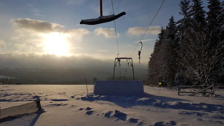 Eine idyllische Winterlandschaft mit schneebedeckten Hügeln und einem Skilift. Die Sonne geht hinter den Bergen unter und taucht die Szene in warmes Licht.