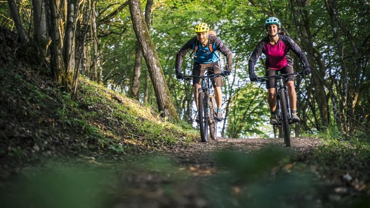 Two cyclists on a forest path, surrounded by green trees. They are wearing helmets and look relaxed.
