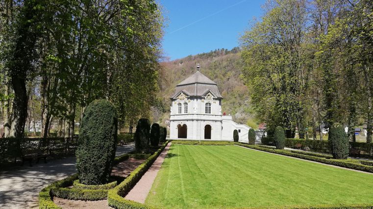 Ein schöner Garten mit gepflegtem Rasen und Symmetrien in der Bepflanzung. Im Hintergrund steht ein elegantes Gebäude umgeben von Bäumen.