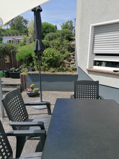 Een gezellige buitenruimte met een tafel en stoelen. Op de achtergrond zijn er planten en een parasol te zien.