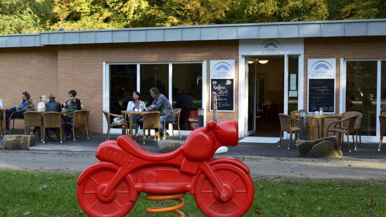 Ein rotes Spielmotorrad steht im Vordergrund. Im Hintergrund sieht man eine Cafeteria mit Gästen, die draußen sitzen.