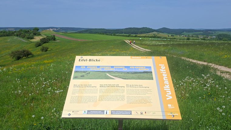 A viewpoint with an information board about the Eifel. The landscape is green and hilly under a clear blue sky.