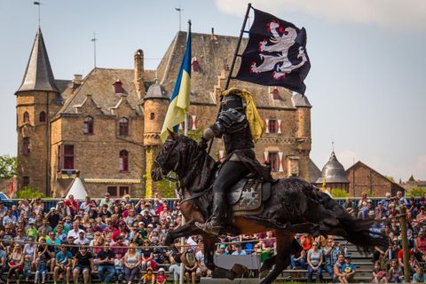 A knight on a horse rides with a flag in hand. In the background, an ancient castle and a large crowd can be seen.