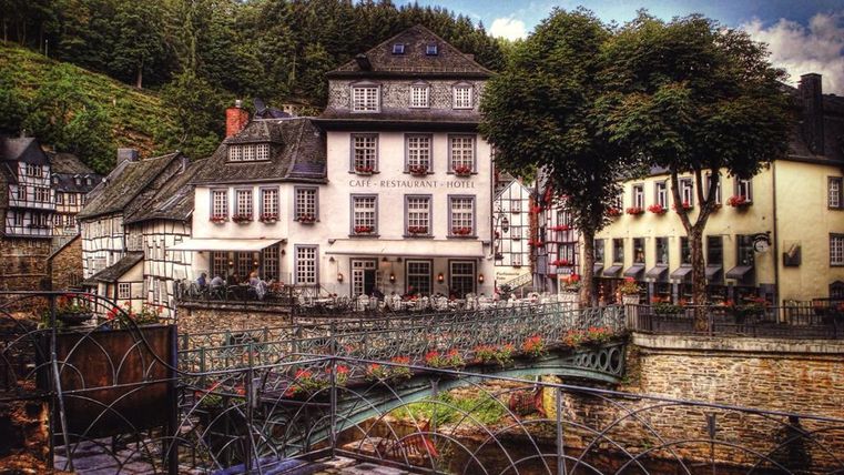 Ein malerisches Gasthaus mit traditioneller Architektur steht an einem kleinen Fluss. Umgeben von Bäumen und Hügeln, lädt der Ort zum Verweilen ein.