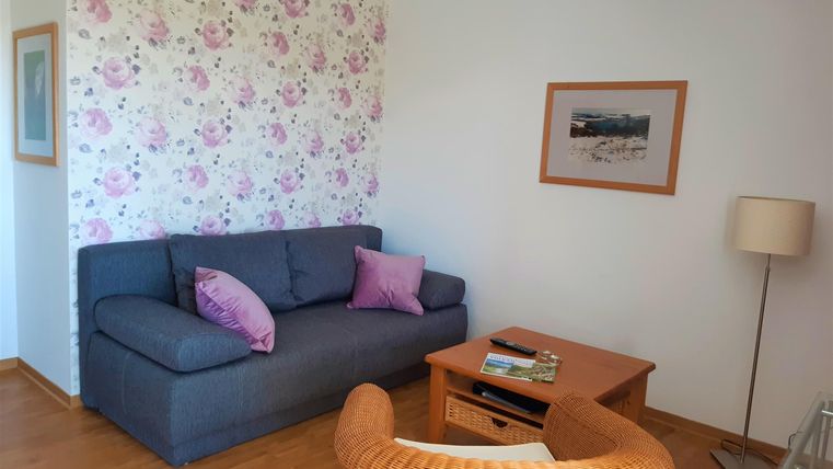 Een gezellige woonkamer met een grijze bank en een rieten stoel. De muren zijn versierd met een bloeiende achtergrondpatroon.