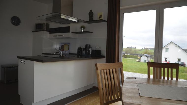 A modern kitchen with a table and chairs. The view of the green landscape is visible through large windows.