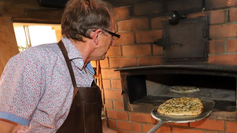 A man takes a freshly baked pizza out of a wood-fired oven. The oven has a rustic brick surface.