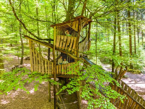 A treehouse made of wood, surrounded by green trees and plenty of foliage. Children are playing in it and enjoying nature.