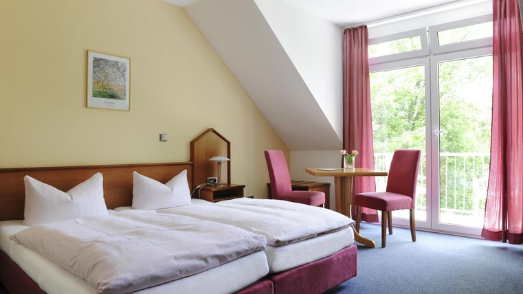 Ein helles Zimmer mit Doppelbett und einem kleinen Esstisch. Große Fenster bieten Blick nach draußen.