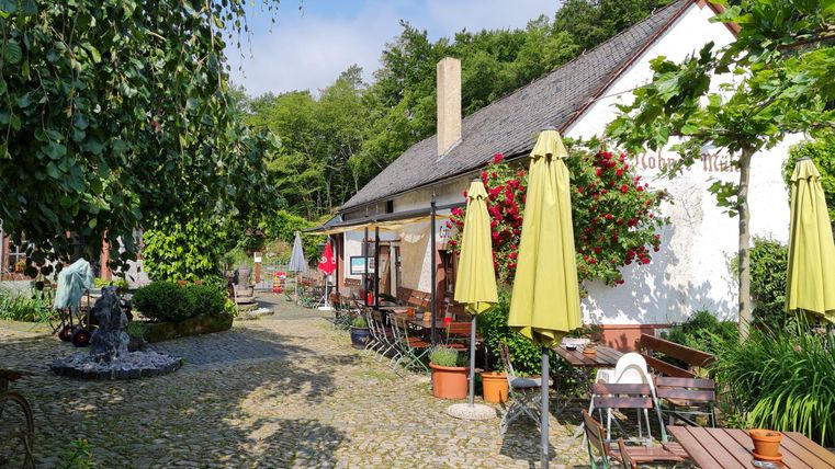 Ein idyllischer Hof mit Tischen, Stühlen und Sonnenschirmen vor einem Gebäude mit der Aufschrift 'Nohner Mühle'.