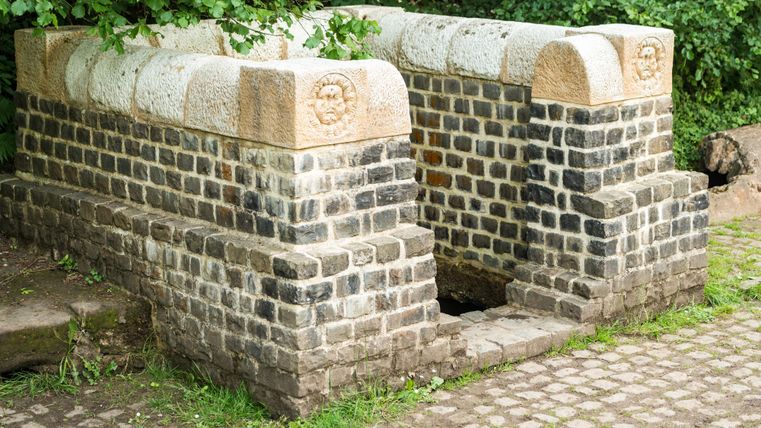 A historical fountain made of stones, surrounded by green grass and trees. The structure has a few decorative stones on top.