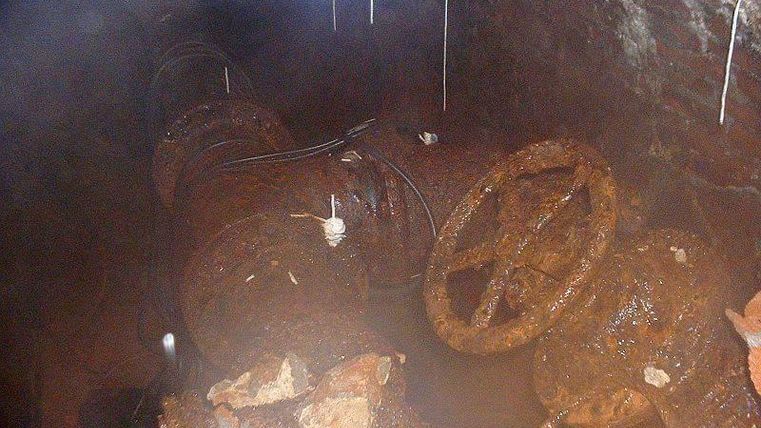 A rusty underground pipe with a valve control unit mounted on top of it. The area looks damp and is surrounded by crumbling bricks.