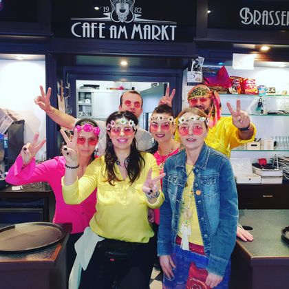 Eine Gruppe von fröhlichen Menschen in bunten Outfits steht in einem Café. Sie tragen Sonnenbrillen und zeigen Frieden-Zeichen.